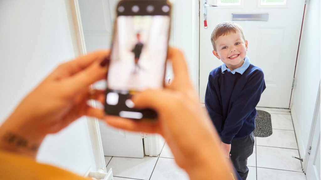 A parent takes a photo of a smiling young boy dressed and ready for school.