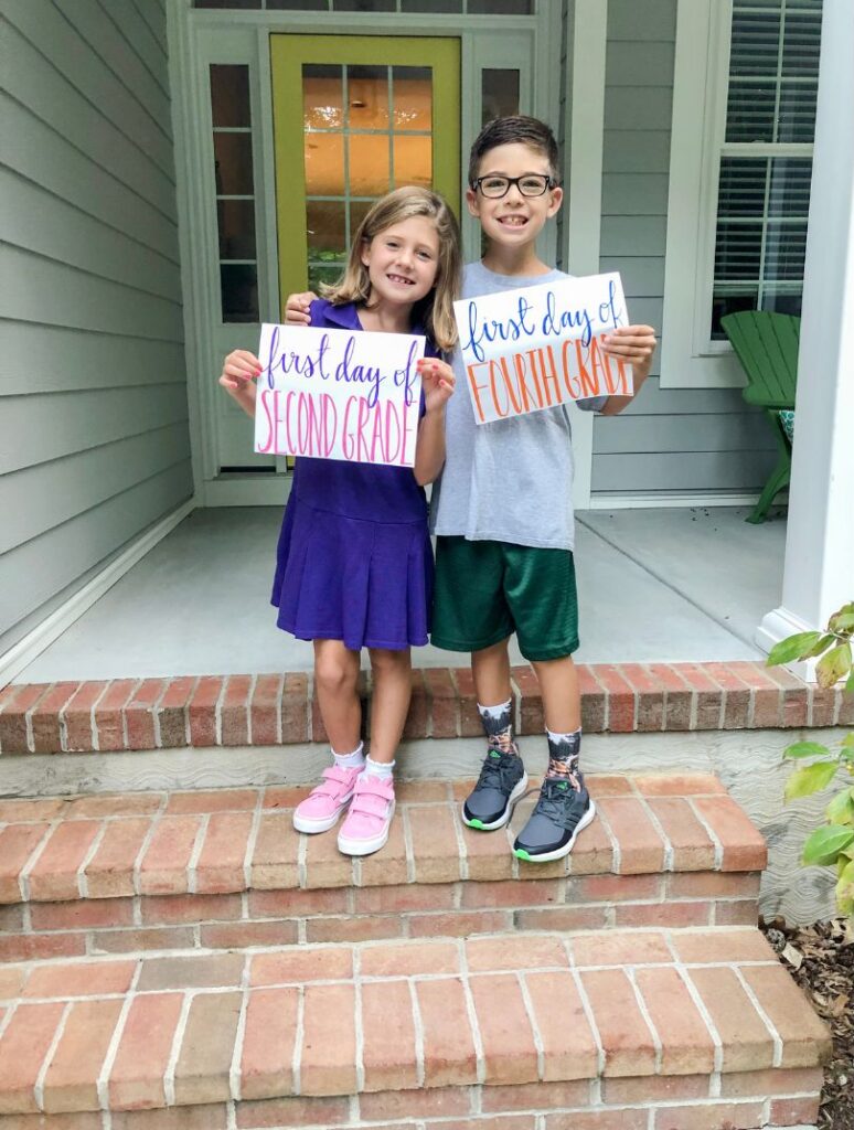 Two children stand on a front porch holding signs for their first days of second and fourth grade.