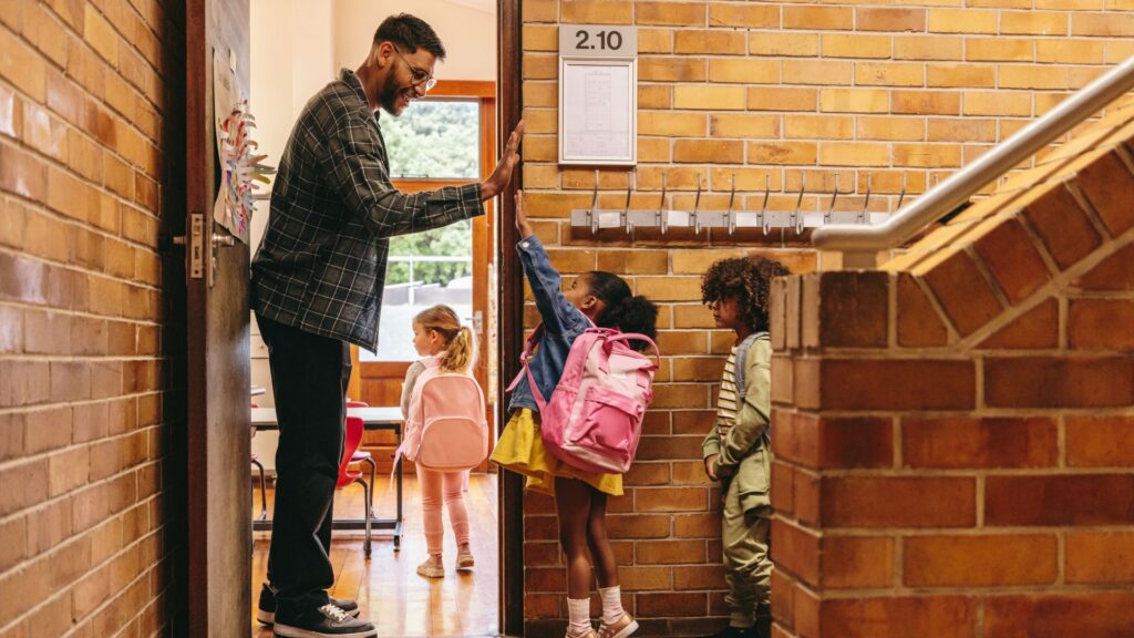 A student wearing a backpack gives her teacher a high five at the classroom door.